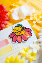 Load image into Gallery viewer, The Sleepy Bee Bamboo Sticker is an eco-friendly accessory featuring an adorable illustration of a drowsy bee. Crafted from 100% sustainable bamboo, this sticker measures approximately 3x3 inches. It is designed to be fade and weather-resistant, making it suitable for various surfaces such as hydro flasks, journals, and tote bags. The strong 3M water-resistant adhesive ensures durability and longevity. Due to the natural variations in bamboo, each sticker may have slight differences in color and appearance.