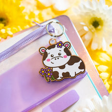 Load image into Gallery viewer, A charming keychain featuring a cow surrounded by a cluster of delicate flowers. The cow has a sweet, cartoonish design with soft features and a peaceful expression, while the colorful flowers add a cheerful and whimsical touch. Made from durable material, this keychain is both cute and practical, perfect for decorating keys, bags, or backpacks