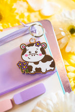 Load image into Gallery viewer, A charming keychain featuring a cow surrounded by a cluster of delicate flowers. The cow has a sweet, cartoonish design with soft features and a peaceful expression, while the colorful flowers add a cheerful and whimsical touch. Made from durable material, this keychain is both cute and practical, perfect for decorating keys, bags, or backpacks