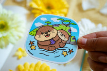 Load image into Gallery viewer, A glossy sticker featuring a cute otter with a playful expression. The otter is either floating on its back or holding a small object, giving off an adorable and cheerful vibe.
Let me know if you’d like any edits! 😊🦦✨