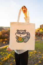 Load image into Gallery viewer, A playful design featuring a mischievous raccoon happily munching on a fresh ear of corn! Printed on a durable, eco-friendly tote bag, perfect for carrying books, groceries, or everyday essentials. A fun and quirky choice for raccoon lovers and foodies!