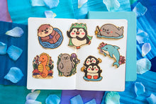 Load image into Gallery viewer, A cute sea otter floating happily on its back, holding a seashell with a cheerful expression. This eco-friendly bamboo sticker features a charming, hand-drawn style, perfect for decorating laptops, water bottles, and journals! 🦦💙✨