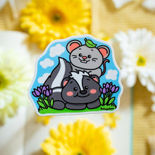 Load image into Gallery viewer, A glossy sticker featuring a cute black-and-white skunk with a fluffy tail and a tiny gray mouse sitting beside it. The two animals appear friendly and curious, creating a charming and heartwarming design.