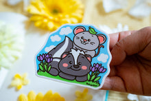 Load image into Gallery viewer, A glossy sticker featuring a cute black-and-white skunk with a fluffy tail and a tiny gray mouse sitting beside it. The two animals appear friendly and curious, creating a charming and heartwarming design.