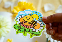 Load image into Gallery viewer, A glossy sticker featuring a vibrant bee hovering over a bright sunflower. The bee is detailed with delicate wings, and the sunflower is in full bloom, adding a cheerful, nature-inspired touch to the sticker