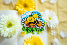 Load image into Gallery viewer, A glossy sticker featuring a vibrant bee hovering over a bright sunflower. The bee is detailed with delicate wings, and the sunflower is in full bloom, adding a cheerful, nature-inspired touch to the sticker