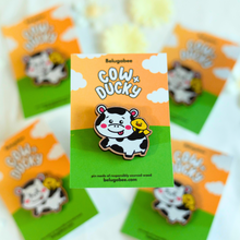 Load image into Gallery viewer, 
A wooden pin featuring an adorable design of a cow and a duck together. The cow, with a friendly and calm expression, is etched with fine details like spots and a subtle wood grain texture. The duck sits cheerfully on the cow's back, adding a playful touch to the design. The natural wood finish gives the pin a rustic, eco-friendly charm, perfect for accessorizing jackets, bags, or hats