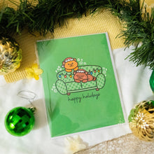 Load image into Gallery viewer, 
A cute and colorful holiday card featuring a cozy scene of a cat lounging on a festive couch. The cat, with a playful and relaxed expression, is surrounded by holiday decorations like string lights, ornaments, and gift boxes. The couch is adorned with cheerful patterns in red, green, and gold, adding a warm and festive vibe. Perfect for spreading holiday cheer with a touch of feline charm!