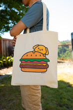 Load image into Gallery viewer, A fun and quirky tote bag featuring an adorable design of a puppy inside a burger. The playful illustration shows a happy puppy popping out between the layers of a juicy burger, complete with lettuce, cheese, and a sesame seed bun. The design is colorful and whimsical, set against a sturdy canvas background, making it perfect for carrying books, groceries, or everyday essentials with a touch of humor and cuteness.