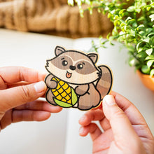 Load image into Gallery viewer, An adorable raccoon with bright, curious eyes, holding a tiny treasure in its paws! This eco-friendly bamboo sticker features soft, earthy tones and a charming design, perfect for adding a touch of woodland whimsy to your water bottle, laptop, or journal. A must-have for raccoon lovers!
