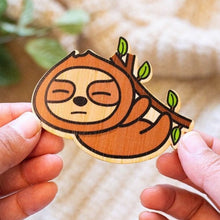 Load image into Gallery viewer, The Sleepy Hanging Sloth Bamboo Sticker is an eco-friendly accessory featuring an adorable illustration of a sloth hanging from a branch, appearing sleepy and content. Crafted from 100% sustainable bamboo, this sticker measures approximately 3x3 inches. It is designed to be fade and weather-resistant, making it suitable for various surfaces such as hydro flasks, journals, and tote bags. The strong 3M water-resistant adhesive ensures durability and longevity. Due to the natural variations in bamboo, each sti