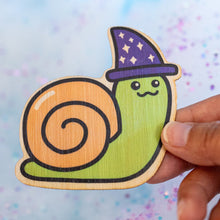 Load image into Gallery viewer, The Wizard Snail Bamboo Sticker is an eco-friendly decorative item featuring a whimsical illustration of a snail adorned with wizard attire. Crafted from 100% sustainable bamboo, this sticker measures approximately 3x3 inches and is designed to be both fade and weather-resistant. Its strong 3M water-resistant adhesive ensures durability across various surfaces, including hydro flasks, journals, and tote bags. This sticker combines environmental consciousness with a touch of fantasy, making it a delightful a