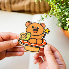 Load image into Gallery viewer, Enter the enchanting realm with our Fairy Bear Bamboo Sticker. This whimsical 3x3-inch sticker features a magical scene of a bear accompanied by a snail, both holding a star wand, seated on a charming white and red mushroom. Meticulously crafted on sustainable bamboo, this eco-friendly sticker adds a touch of fantasy to any surface. Elevate your style with the Fairy Bear Bamboo Sticker – a perfect blend of charm and environmental consciousness. 🧚🐻🍄 #BambooSticker #FairyBear #EnchantedDecor
