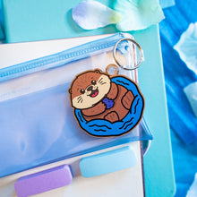 Load image into Gallery viewer, A cute wooden keychain featuring a playful otter floating on its back, holding a small object. This eco-friendly keychain has a charming, hand-carved design, perfect for adding a touch of oceanic charm to your keys or bag! 🦦🌊✨
