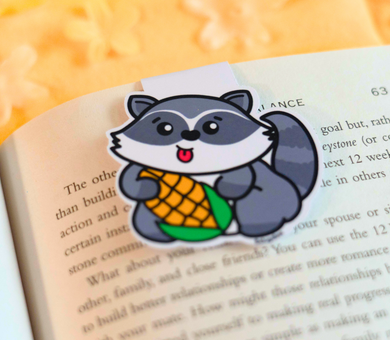 A fun magnetic bookmark featuring a mischievous raccoon holding a piece of corn with a cheeky grin. This eco-friendly bookmark has a playful, hand-drawn style, perfect for adding a touch of whimsy to your reading! 🦝🌽📖✨