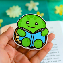 Load image into Gallery viewer, A glossy sticker featuring a turtle reading a book, available on Redbubble
