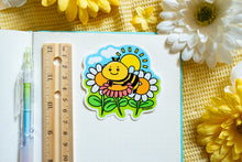 Load image into Gallery viewer, A glossy sticker featuring a vibrant bee hovering over a bright sunflower. The bee is detailed with delicate wings, and the sunflower is in full bloom, adding a cheerful, nature-inspired touch to the sticker
