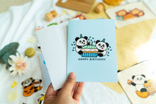 Load image into Gallery viewer, A charming birthday card featuring adorable pandas, each with a joyful expression, surrounding a delicious-looking cupcake topped with colorful frosting and a candle. The card includes the message &quot;Happy Birthday&quot; in playful, vibrant lettering, adding a festive touch to the celebration
