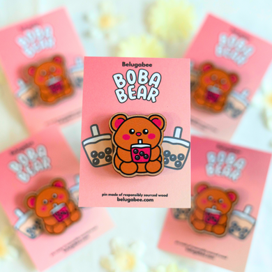 A wooden pin shaped like a bear holding a cup of boba tea. The bear has a cute, cartoonish design with rounded features and a friendly expression. The cup of boba tea is detailed with tiny tapioca pearls at the bottom, and the bear appears to be happily sipping through a straw. The wood has a natural grain, adding a rustic charm to the design