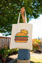 Load image into Gallery viewer, A fun and quirky tote bag featuring an adorable design of a puppy inside a burger. The playful illustration shows a happy puppy popping out between the layers of a juicy burger, complete with lettuce, cheese, and a sesame seed bun. The design is colorful and whimsical, set against a sturdy canvas background, making it perfect for carrying books, groceries, or everyday essentials with a touch of humor and cuteness.
